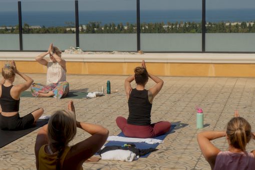 Outdoor yoga session on a rooftop terrace by the sea; people sit on mats, practicing poses as a guided instructor leads.