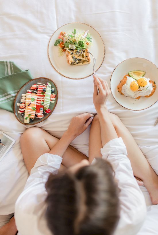 A person sits cross‑legged on a bed with three plates of food: sushi skewers, salad, and a dessert or fruit dish, all seen from above.