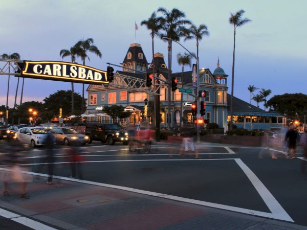 Carlsbad landmark at dusk: a historic train station building, palm trees, and a lit sign over a busy beachfront street with people and cars moving.