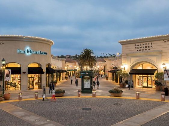 A sunny outdoor shopping plaza with Mediterranean-style storefronts, cobblestone paths, palm trees, and shoppers strolling along.