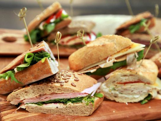 Assorted deli sandwiches with herbs and toothpick skewers on a wooden board, cut lunches ready to serve.