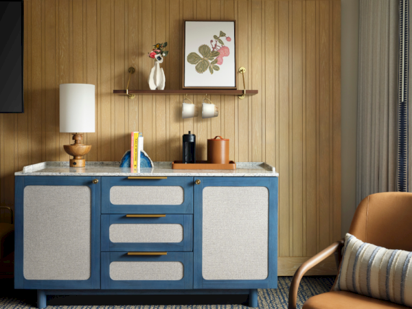A mid-century blue cabinet with cream doors flanked by a lamp, bookshelf items, and a brown leather chair in a cozy wooden nook.