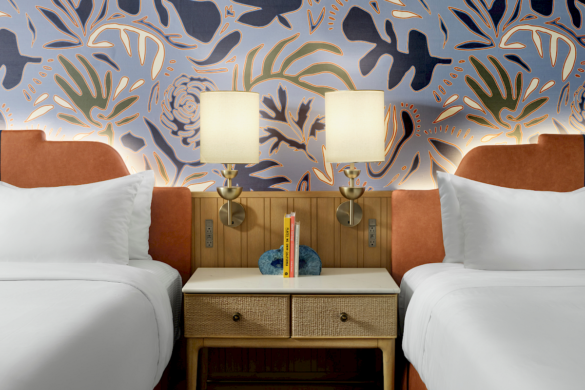 Two twin beds with white linens flank a wooden nightstand between them; matching lamps glow against bold blue floral wallpaper, cozy vibe.