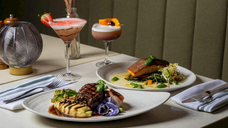 Two fancy dishes with steak and fish, plus two cocktails on a chic table&mdash;gourmet plates, garnishes, and a cozy dining vibe.